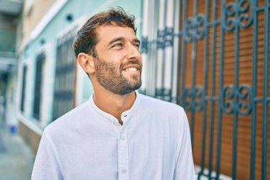 Güneşli bir günde günlük beyaz gömlek giyen sakallı yakışıklı adam dışarıda mutlu bir şekilde gülümsüyor.