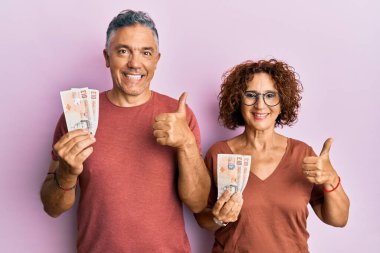 Güzel orta yaşlı çift ellerinde 10 pound tutuyor mutlu ve pozitif gülümseyen banknotlar, başparmak yukarı mükemmel ve onay işareti yapıyor 