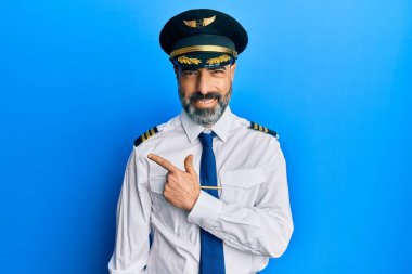 Orta yaşlı, sakallı, gri saçlı, uçak pilotu üniforması giyen neşeli, yüzünde mutlu ve doğal bir ifadeyle el ve parmak uçlarını gösteren bir gülümsemesi olan bir adam. 