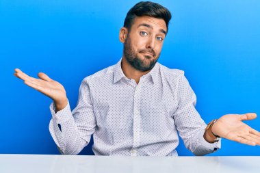 Masada oturan, iş kıyafetleri giymiş, elleri ve kolları havada, kafası karışmış yakışıklı İspanyol bir adam. şüphe kavramı. 