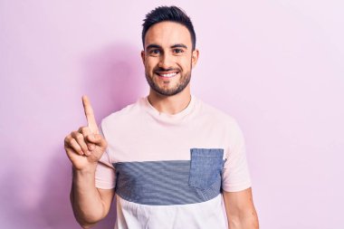 Sakallı, sıradan tişörtlü, pembe arka planda duran, bir fikir veya soruyla gülümseyen, mutlu yüzlü, genç yakışıklı bir adam.