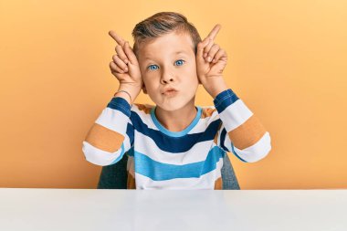 Kafkasyalı sevimli bir çocuk, günlük giysiler içinde masada oturmuş, boynuzlarını başlarının üzerinde gezdirirken komik jestler yapıyor. 