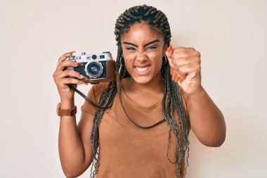 Saç örgülü, elinde eski model fotoğraf makinesi olan Afrikalı genç bir kadın öfkeyle bağırıyor, öfkeyle bağırıyor ve el kaldırıyor. 