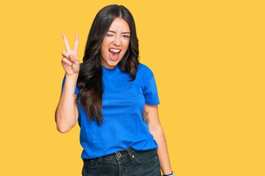 Güzel esmer, sıradan kıyafetler giyen mutlu yüzüyle gülümseyen ve parmaklarıyla zafer işareti yapan genç bir kadın. İki numara.. 