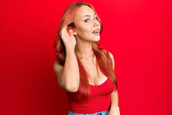 若い美しい赤毛の女性は 耳を傾け 噂やゴシップに耳を傾けながら手で笑みを浮かべて赤い背景にカジュアルな服を着ています 聴覚障害の概念 — ストック写真