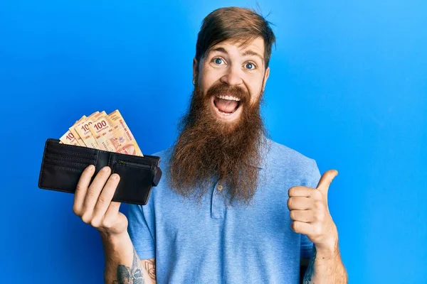 額を指しているメキシコのペソで財布を保持している長いひげを持つ赤毛の男まで笑顔で幸せなオープン口 — ストック写真