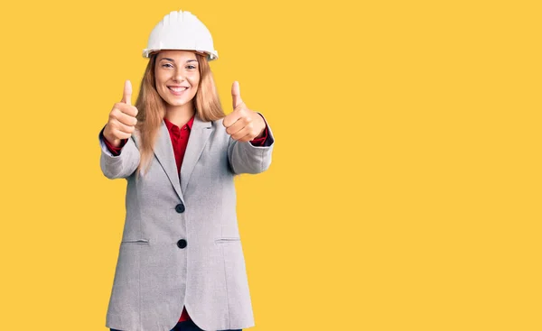 漂亮的年轻女子戴着建筑师的硬礼帽 赞成用手做积极的手势 微笑着竖起大拇指 为成功感到高兴 优胜手势 — 图库照片