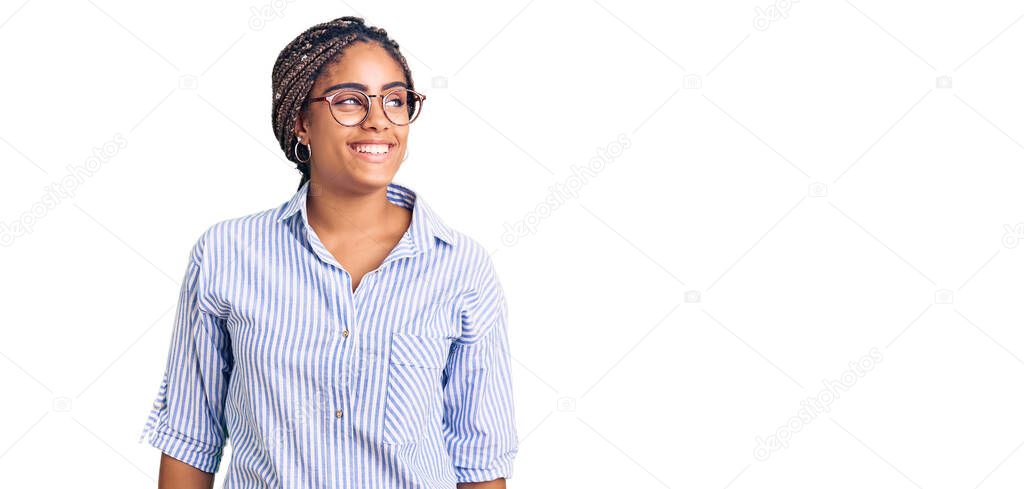 Young african american woman with braids wearing casual clothes and glasses looking away to side with smile on face, natural expression. laughing confident. 