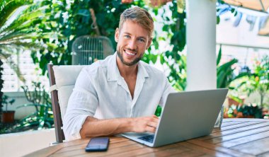 Terasta oturan yakışıklı beyaz adam bilgisayar laptopu kullanarak evden çalışıyor.