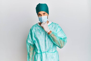 Cerrah üniforması giyen ve tıbbi maske takan genç bir adam kameraya kollarını kavuşturup gülümseyerek ve çenesini kaldırarak bakıyor. Olumlu düşünmek. 