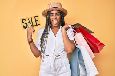 Saç örgülü, alışveriş torbaları ve bayrak taşıyan genç Afro-Amerikalı kadın komik bir ifadeyle dilini dışarı çıkartıyor.. 