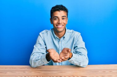 Genç, yakışıklı, Afro-Amerikan bir adam, günlük giysiler giymiş, ellerini masaya koyup gülümsüyor, el ele tutuşuyor ya da jest yapıyor. Bekleme ve koruma 