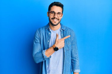 Gündelik kıyafetler ve gözlük takan genç İspanyol adam gülümsüyor. Elleri ve parmakları yana bakıyor. 