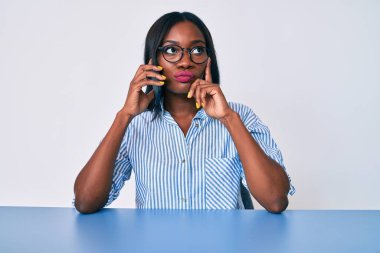 Genç Afro-Amerikan kadın akıllı telefonuyla konuşuyor masada oturuyor ciddi bir yüz ifadesi var eli çeneli soruyu düşünüyor, kafa karıştırıcı bir fikir hakkında düşünceli. 