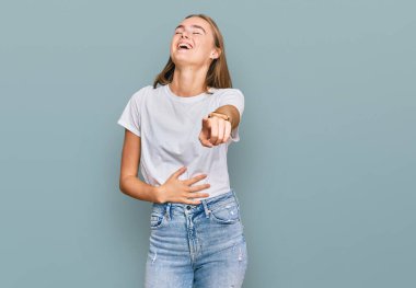 Güzel sarışın kadın, günlük beyaz tişört giyip sana gülüyor, kamerayı işaret ediyor, vücudunu gösteriyor, utanç ifadesi var. 