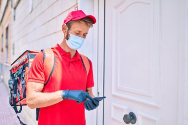 Beyaz postacı kırmızı üniforma ve teslimat çantası giyiyor ve akıllı telefon kullanarak koronavirüs güvenlik maskesi takıyor.