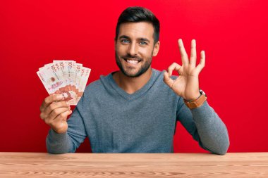 Handsome hispanic man holding colombian pesos doing ok sign with fingers, smiling friendly gesturing excellent symbol 