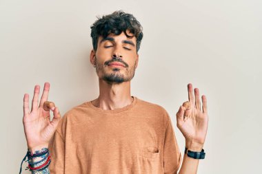 Günlük kıyafetler giyen genç İspanyol adam rahatla ve gözleri kapalı gülümse parmaklarıyla meditasyon hareketi yapıyor. yoga konsepti. 