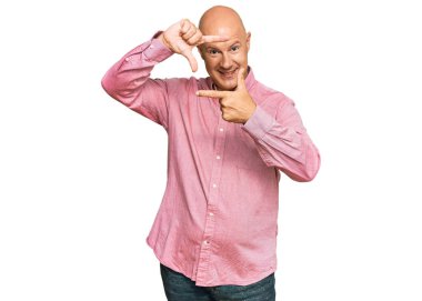 Orta yaşlı, kel, sıradan kıyafetler giyen, gülümseyen, elleri ve parmakları mutlu yüzlü bir çerçeve yapan bir adam. yaratıcılık ve fotoğrafçılık kavramı. 