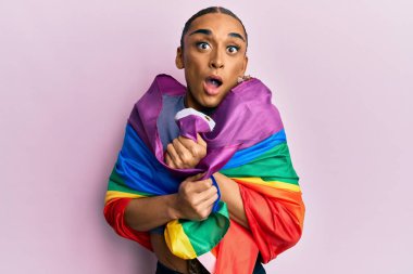 Makyaj yapmış, uzun saçlı, gökkuşağı lgbtq bayrağına sarılı İspanyol bir adam. Şaşırmış ve şaşırmış bir ifade, korku ve heyecanlı bir yüz.. 