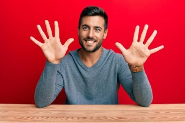 Yakışıklı İspanyol bir adam gündelik tarzda giyinmiş masada oturuyor ve 10 numaralı parmaklarını göstererek gülümsüyor. Kendinden emin ve mutlu.. 