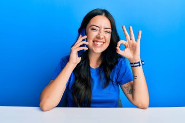 Güzel esmer genç bir kadın akıllı telefonda konuşuyor parmaklarıyla imza atıyor, gülümsüyor ve mükemmel bir sembol oluşturuyor. 