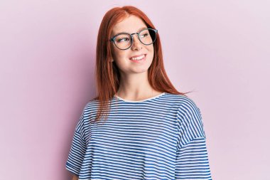 Genç kızıl saçlı, günlük kıyafetler ve gözlük takan rahat bir görünüm takınan, doğal yüzlü ve kendine güvenen bir gülümseme takınan kız.. 