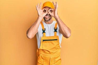 Sarı arka plan üzerinde becerikli üniforması olan genç yakışıklı bir adam dürbün gibi jest yapıyor, dili dışarı çıkarıyor, gözleri parmaklarından bakıyor. çılgın ifade. 