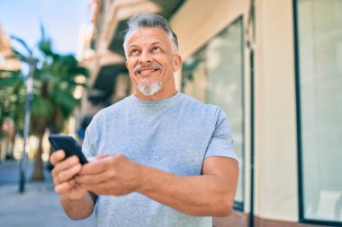 Orta yaşlı, ispanyol, kır saçlı, mutlu gülümseyen adam şehirde akıllı telefon kullanıyor..