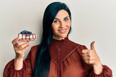 Young caucasian woman holding cake slice smiling happy and positive, thumb up doing excellent and approval sign 