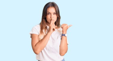 Gündelik beyaz tişört giyen güzel beyaz kadın sessiz olmak istiyor. Parmakları dudaklarında ve elleri yandan işaret ediyor. sessizlik ve gizli konsept. 