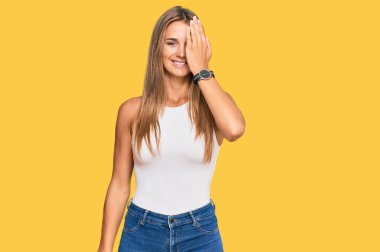 Kolsuz tişörtlü, sıradan giyinen genç sarışın kadın bir gözünü eliyle kapatıyor, yüzünde kendine güvenen bir gülümseme ve sürpriz bir duygu var.. 