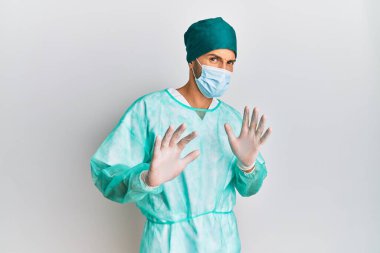 Cerrah üniforması giyen genç yakışıklı bir adam ve tıbbi maske takan tiksindirici bir ifade, tiksinti tepkisi yüzünden tiksinti ve korku dolu bir ifade. Eller havada. 