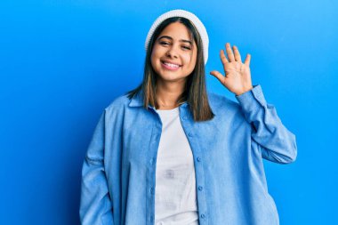 Tatlı yün şapkalı genç Latin kadın merhaba diyor mutlu ve gülümsüyor, dostça karşılama hareketi yapıyor. 