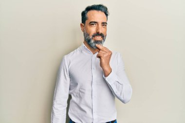 Orta yaşlı, sakallı, gri saçlı, sıradan beyaz tişörtlü kameraya kollarını çapraz yapıp çenesine dokunarak gülümseyen bir adam. Olumlu düşünmek. 