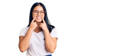 Genç ve güzel Asyalı kız günlük kıyafetler ve gözlük takıyor ağzı açık gülümsüyor, parmakları işaret ediyor ve zorla neşeli bir gülümseme sergiliyor. 