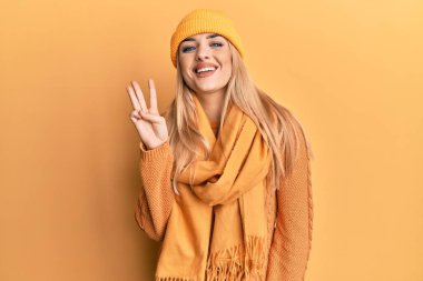 Beyaz kadın yün kazak ve kep giyer ve üç numaralı parmaklarıyla işaret eder, gülümser ve mutlu olur.. 