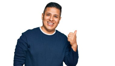 Gündelik kıyafetler giyen, gülümseyen, gülümseyen ve başparmağıyla yan tarafı gösteren genç Latin adam.. 