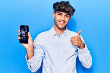 Kırık akıllı telefon tutan genç İspanyol adam çatlak ekranı gösteriyor mutlu ve pozitif gülümsüyor, başparmağıyla mükemmel ve onay işareti yapıyor. 