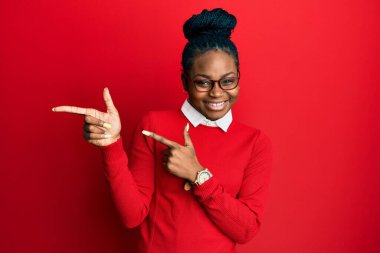 Genç Afrikalı Amerikalı kadın günlük kıyafetler ve gözlük takıyor gülümsüyor ve kameraya bakıyor iki eliyle ve parmaklarıyla yan tarafa bakıyor.. 