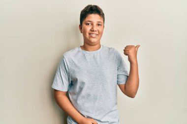 Gündelik gri tişört giyen, gülümseyen, gülümseyen ve baş parmağıyla yan tarafı gösteren genç İspanyol çocuk.. 