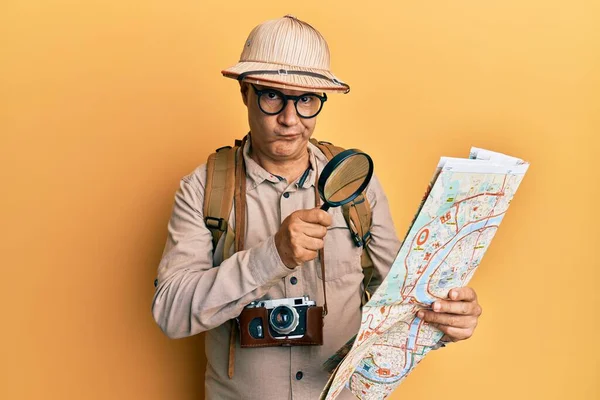 地図上で虫眼鏡を保持探検家の帽子をかぶっている中年のはげ男は懐疑的で緊張しており 問題のために動揺して眉をひそめています 否定的な人 — ストック写真