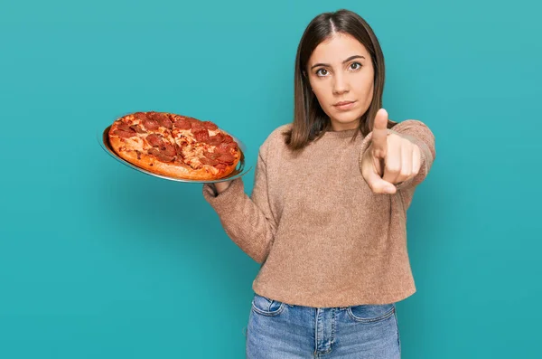 年轻美丽的女人拿着意大利披萨 用手指着相机和你 自信地摆出一副严肃的姿势 — 图库照片