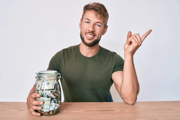 Joven Hombre Caucásico Sosteniendo Frasco Con Ahorros Sentado Mesa Sonriendo —  Fotos de Stock