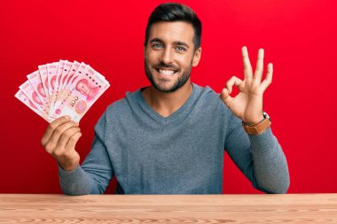 Yakışıklı İspanyol adam Yuan Çin banknotlarını tutuyor tamam işareti yapıyor parmaklarıyla, gülümseyerek mükemmel bir sembol oluşturuyor. 