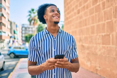 Genç Afrikalı Amerikalı adam şehirde akıllı telefon kullanarak mutlu bir şekilde gülümsüyor..