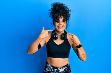 Spor kıyafetleri giyen, kulaklık takan, gülümseyen, parmaklarıyla ve ağzıyla işaret eden İspanyol bir kadın. diş sağlığı kavramı. 