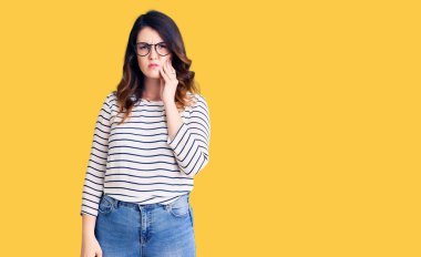 Güzel, genç, esmer, günlük kıyafetler giyen ve gözlüklü bir kadın diş ağrısı ya da diş hastalığından dolayı ağızlarına acı dolu bir ifadeyle dokunuyor. dişçi 