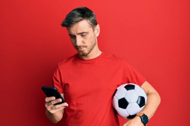 Futbol topu tutan yakışıklı beyaz adam akıllı telefona bakıyor. Yüzünde ciddi bir ifadeyle rahatlamış. Basit ve doğal bir şekilde kameraya bakıyorsun.. 