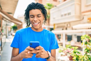 Genç Afrikalı Amerikalı adam şehrin caddesinde akıllı telefon kullanarak mutlu gülümsüyor..